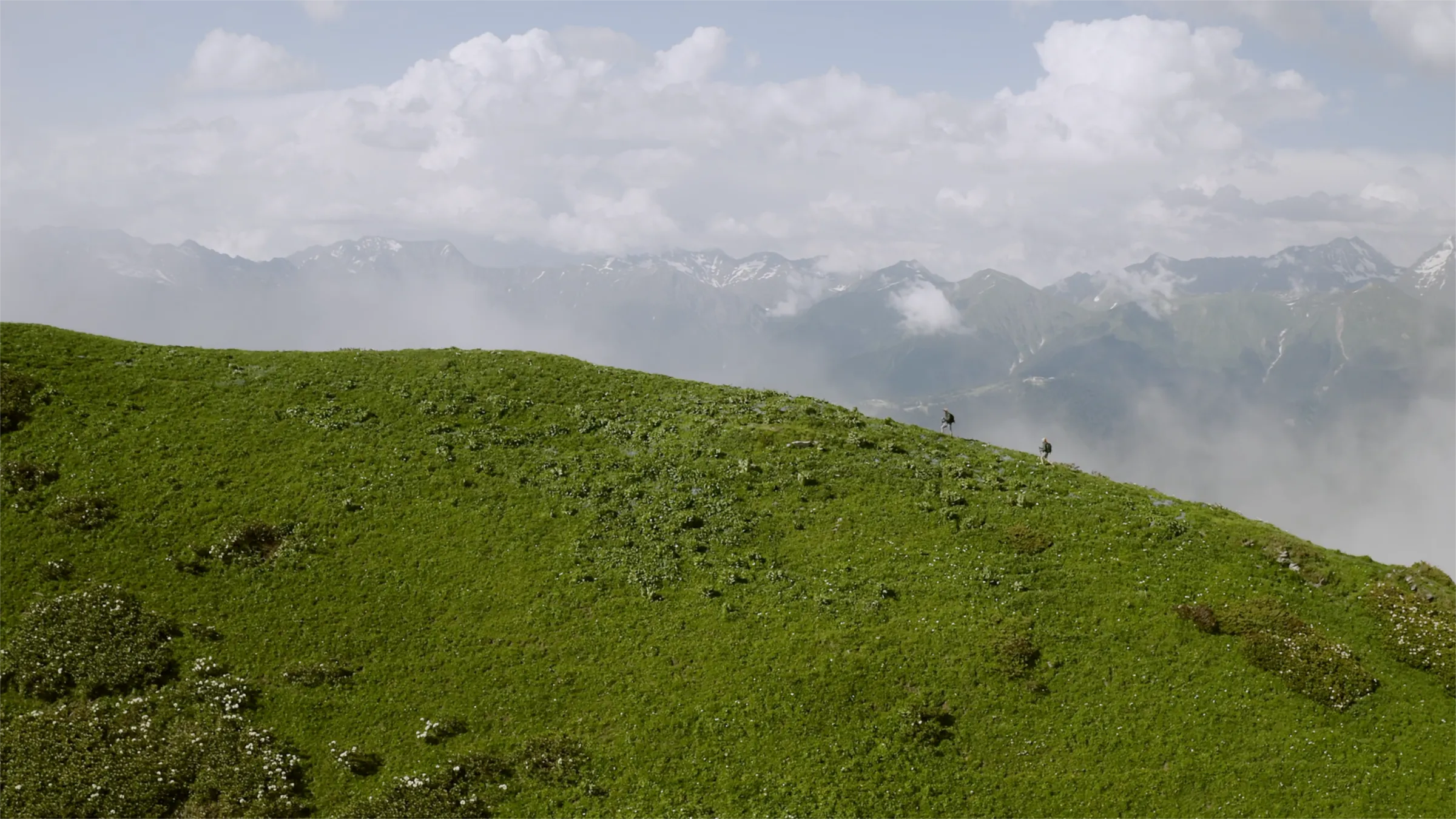 Two hikers walking across a lush green mountain ridge with misty mountains in the background under a cloudy sky.