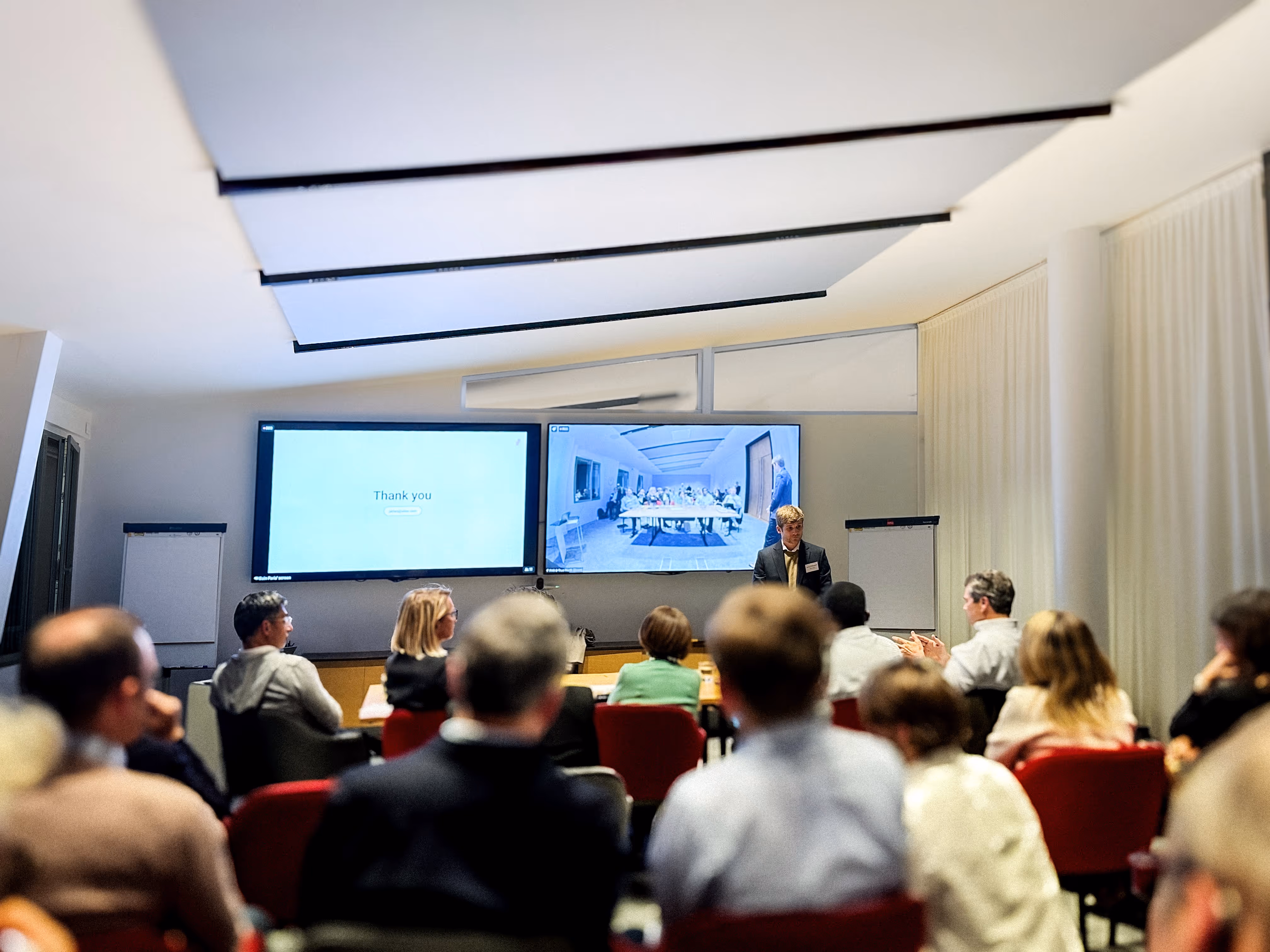 Conference room with attendees facing two large screens, one displaying 'Thank you' and the other showing a video call with a group of people.