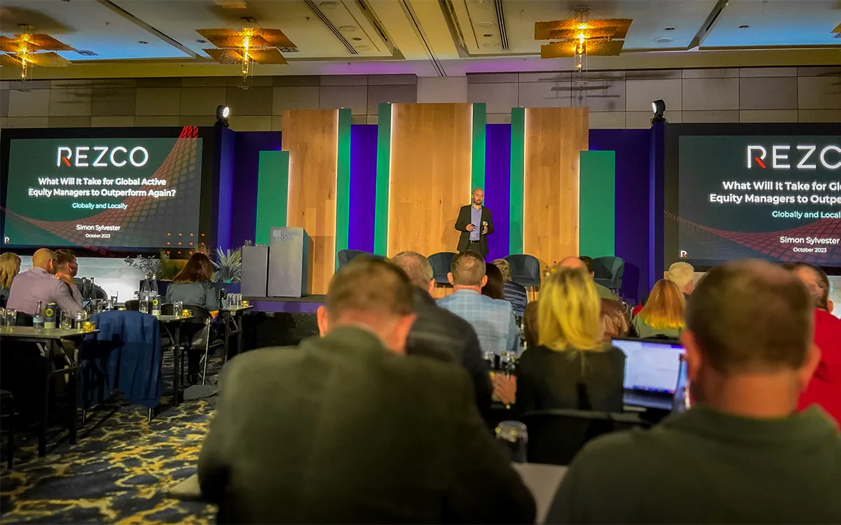 Speaker presenting on stage at REZCO event with audience seated and two large screens showing the presentation title.