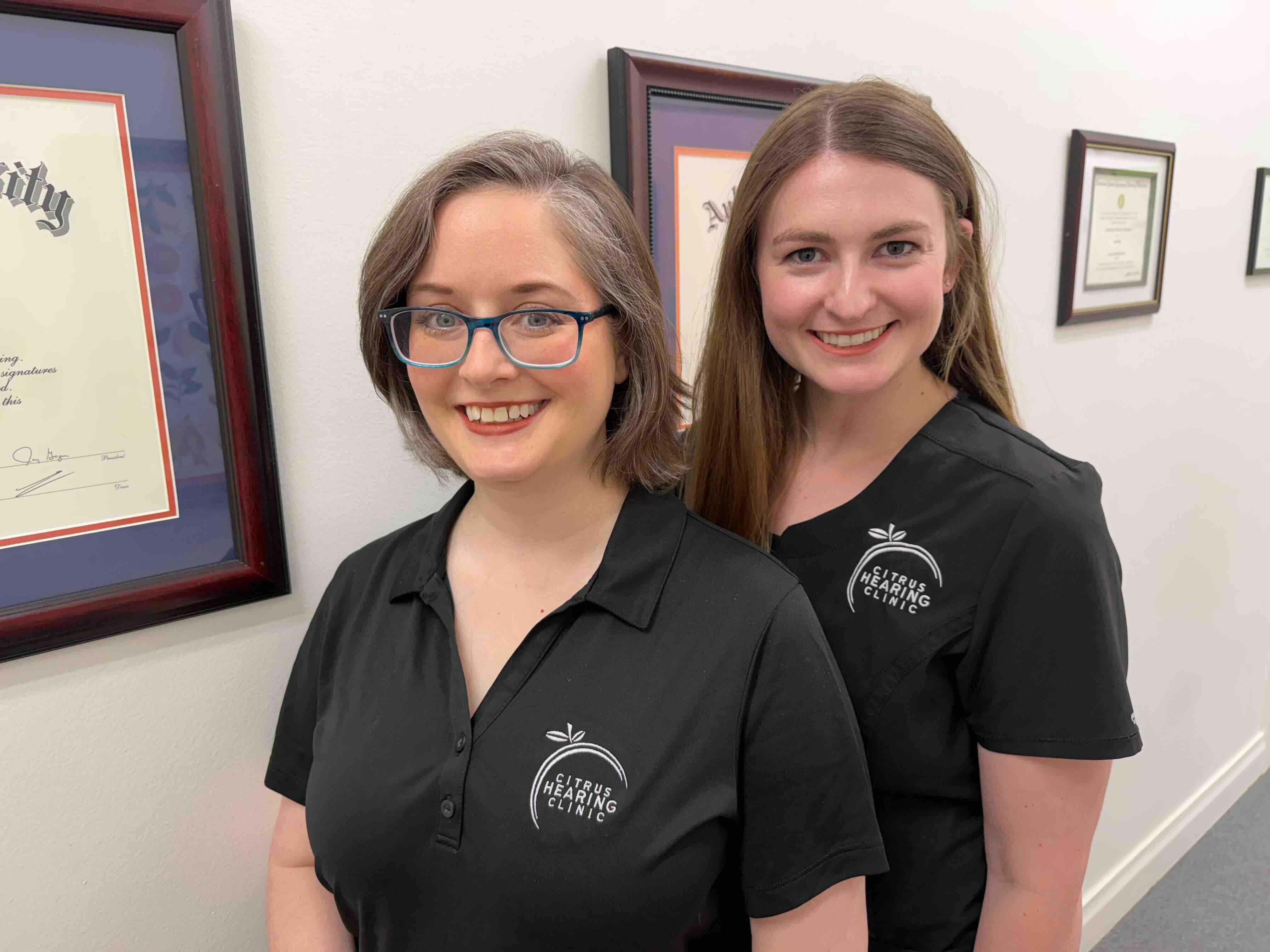 Dr Laura and Dr Teresa wearing white lab coats outdoors, one in a bright pink top and glasses, the other in a blue top with glasses.