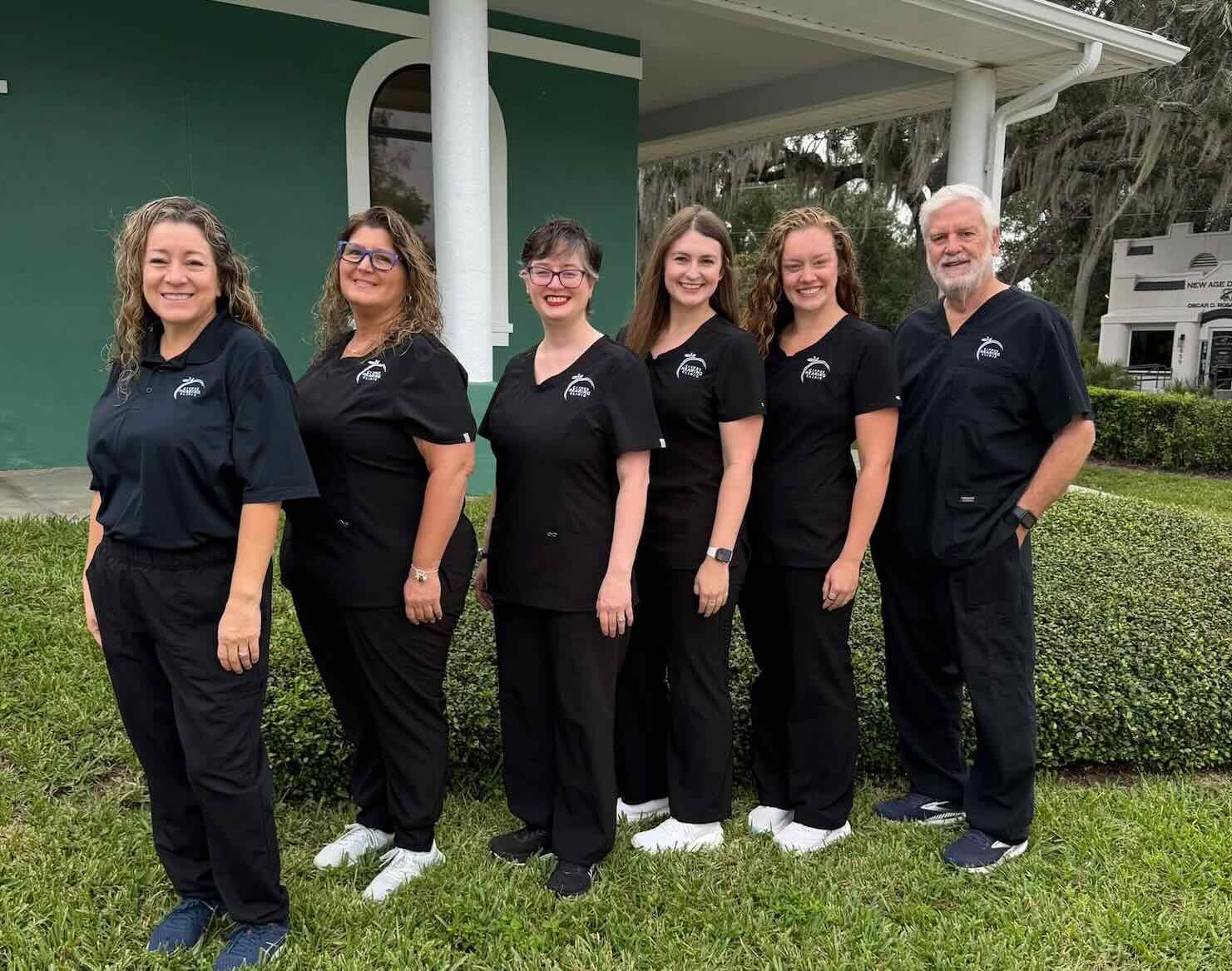 Eight medical professionals standing outdoors in a line, wearing black scrubs in front of a green building with white columns.