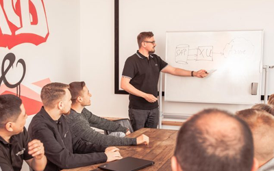 Mann hält Vortrag vor einem Whiteboard und mehrere Personen sitzen um einen Tisch in einem Besprechungsraum.