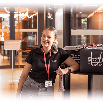 Lächelnde Frau mit Firmenpoloshirt und Namensschild lehnt an einem Tisch mit Community Bags in einem modernen Innenraum.