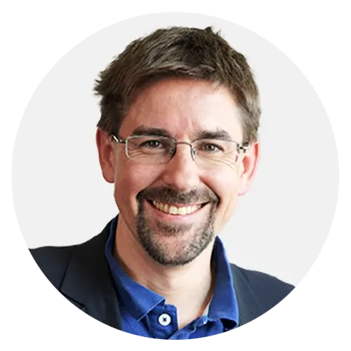 Ein Mann mit braunen Haaren, Brille und Bart lächelt, trägt ein blaues Hemd und eine dunkle Jacke.