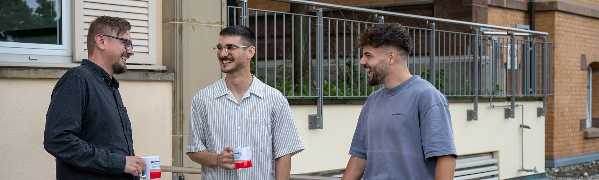 Drei Männer mit Tassen stehen zusammen und unterhalten sich und lachen.
