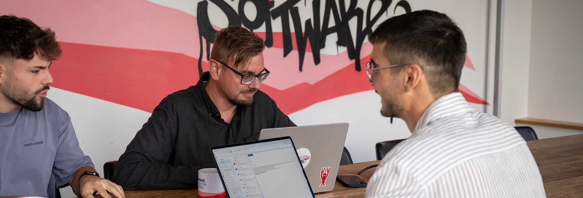Drei Männer sitzen an einem Holztisch mit Laptops und diskutieren in einem Büro mit roter Wandgrafik im Hintergrund.