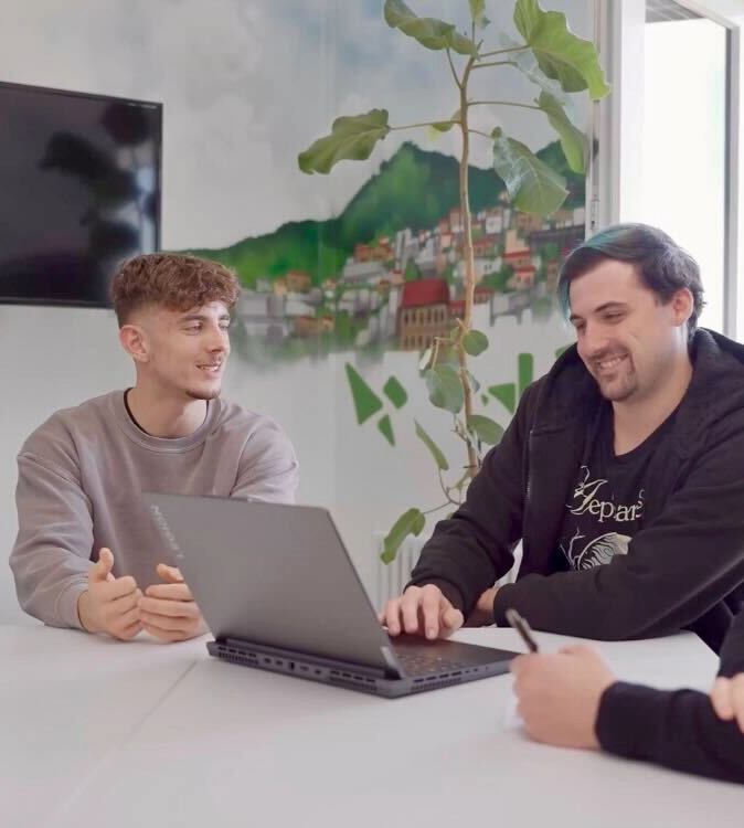 Zwei Männer sitzen an einem Tisch und schauen in einen Laptop.