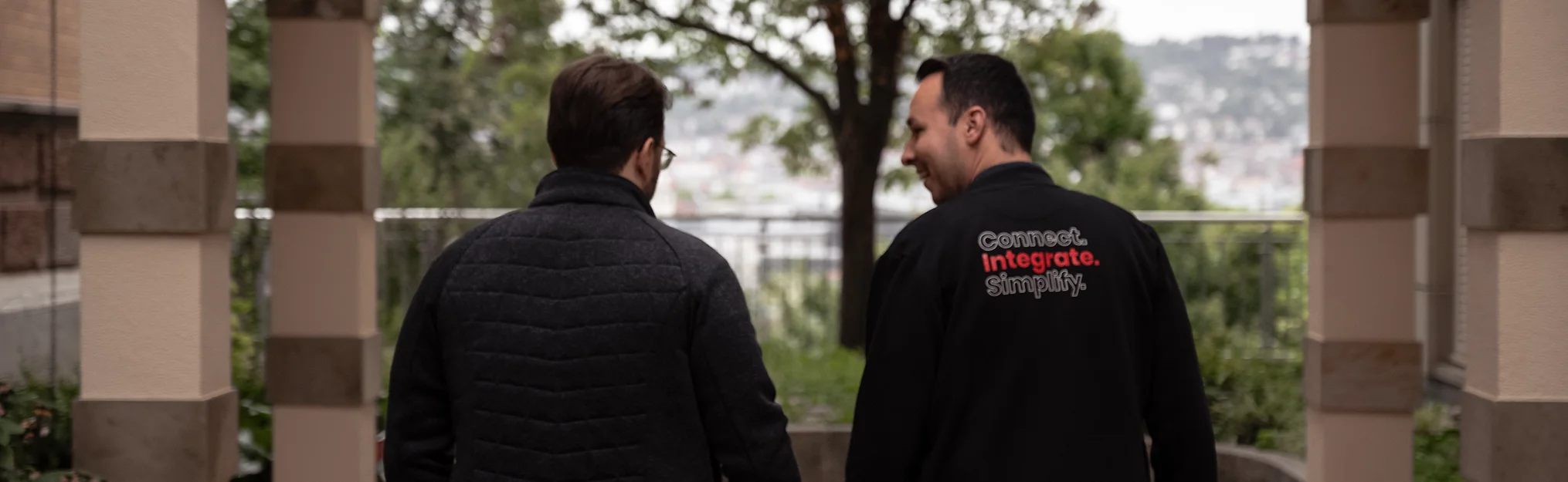 Zwei Männer in Jacken, von hinten aufgenommen, einer trägt eine Jacke mit dem Text ‚Connect. Integrate. Simplify.‘ auf dem Rücken.