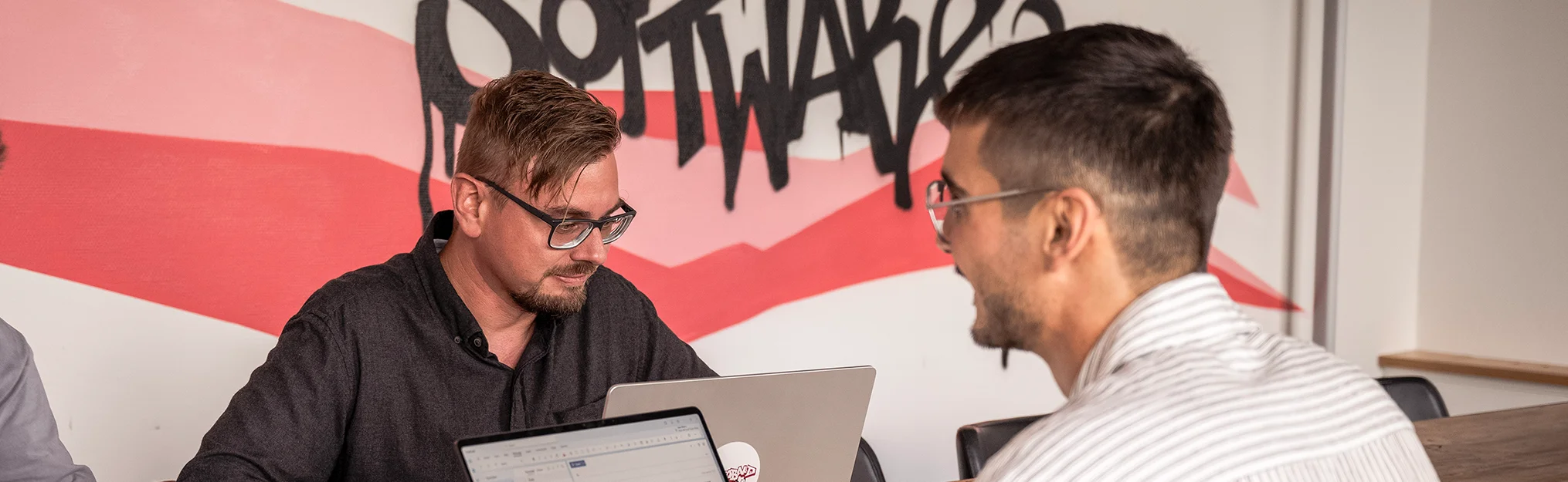 Zwei Männer mit Brillen arbeiten an Laptops vor einer Wand mit grafischem Schriftzug.