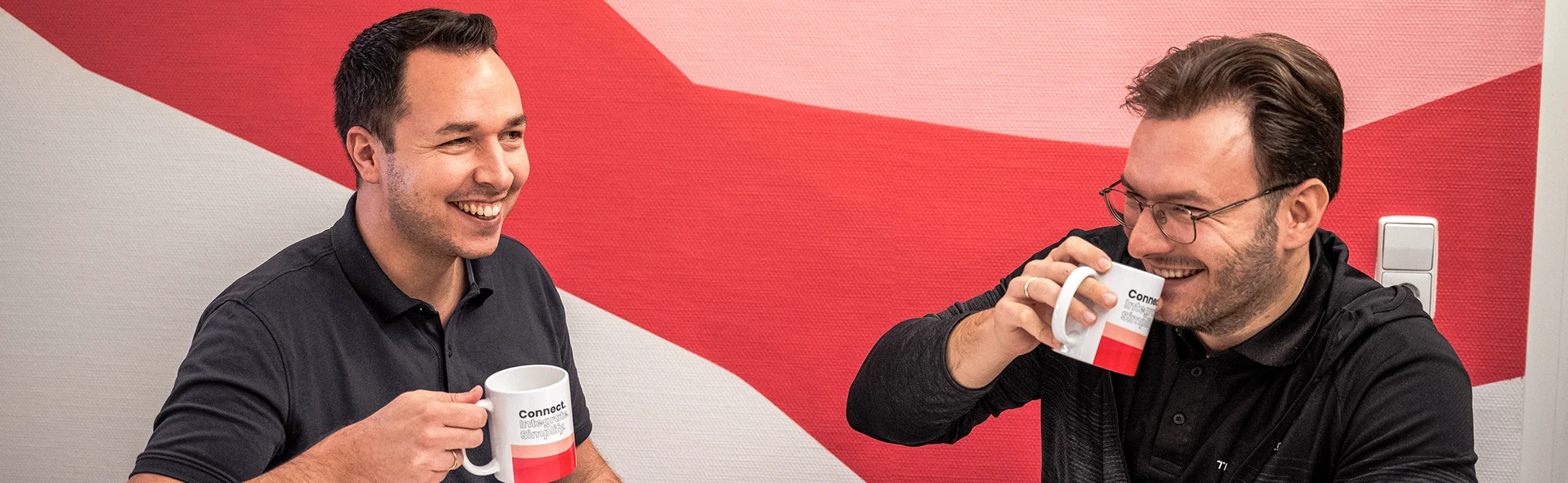 Zwei Männer in schwarzem Polohemd sitzen lachend zusammen und trinken Kaffee aus weißen Tassen mit rotem Design vor einer rot-weißen Wand.