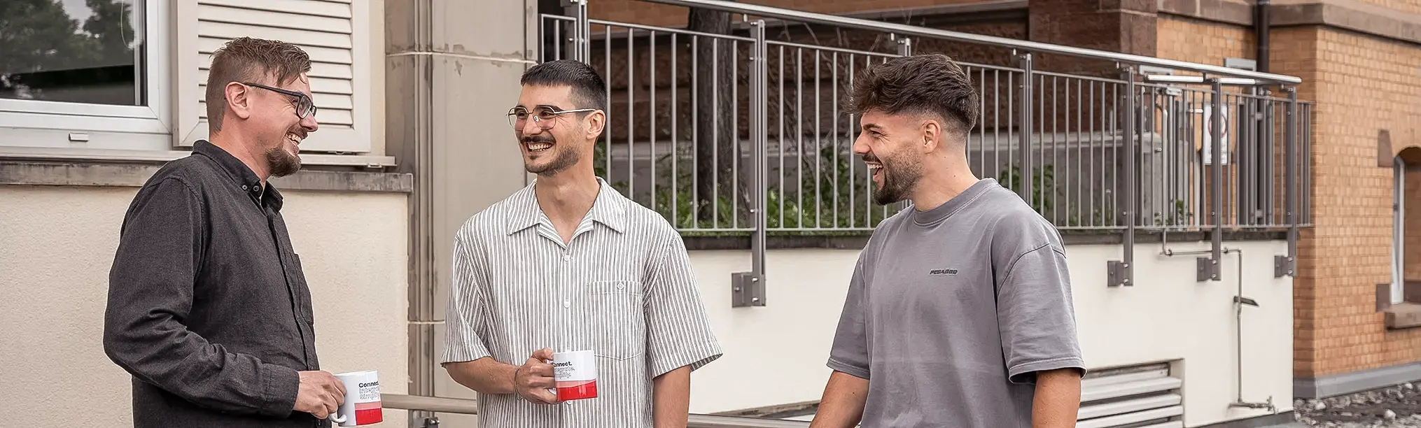 Drei Männer lachen und unterhalten sich draußen bei einer Kaffeepause, zwei halten weiße Tassen mit rotem Boden.