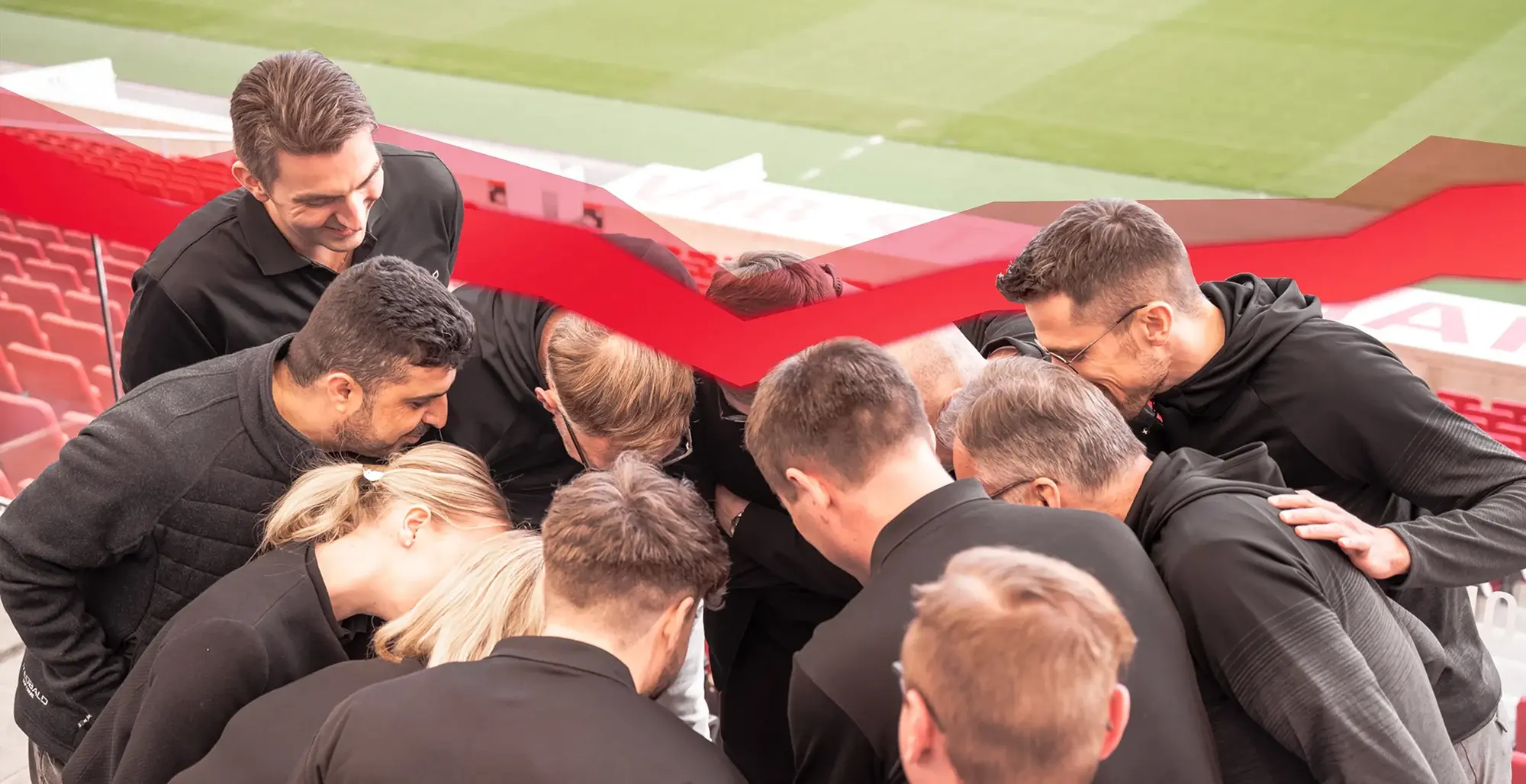 Ein Team von elf Personen steht eng zusammen und beugt sich über etwas in der Mitte, im Hintergrund sind rote Stadiontribünen und ein grünes Spielfeld sichtbar.