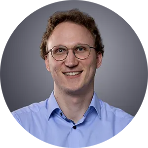 Porträt eines lächelnden Mannes mit lockigem Haar, Brille und blauem Hemd vor grauem Hintergrund.