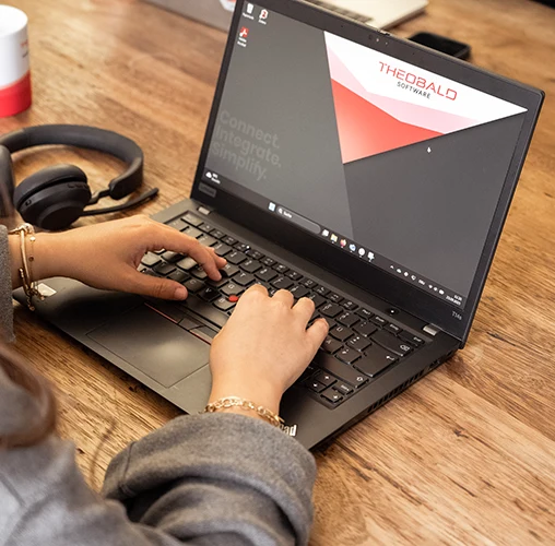 Person typing on a laptop with Theobald Software website on screen, headphones on wooden table.