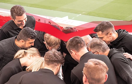 A group of people dressed in black are standing close together in a soccer stadium, bent over something.