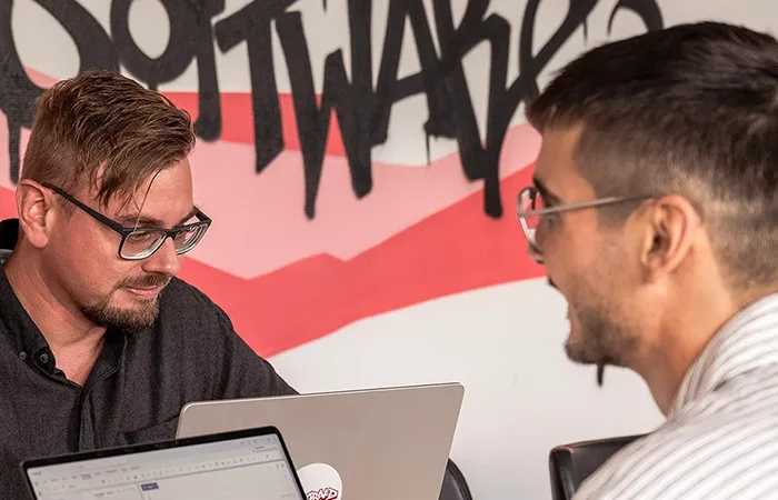 Zwei Männer mit Brillen arbeiten an Laptops vor einer Wand mit grafischem Schriftzug.
