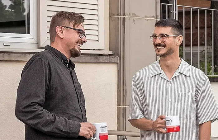 Drei Männer lachen und unterhalten sich draußen bei einer Kaffeepause, zwei halten weiße Tassen mit rotem Boden.