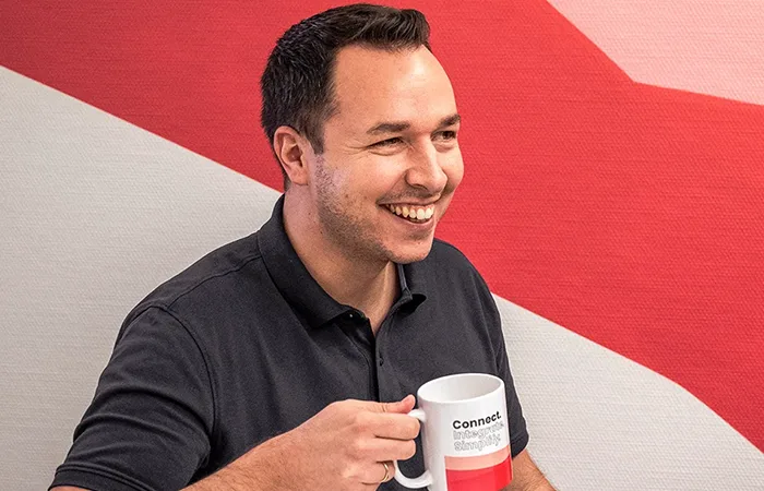 Zwei Männer lachen und trinken Kaffee aus weißen Tassen vor einer rot-rosa gemusterten Wand.