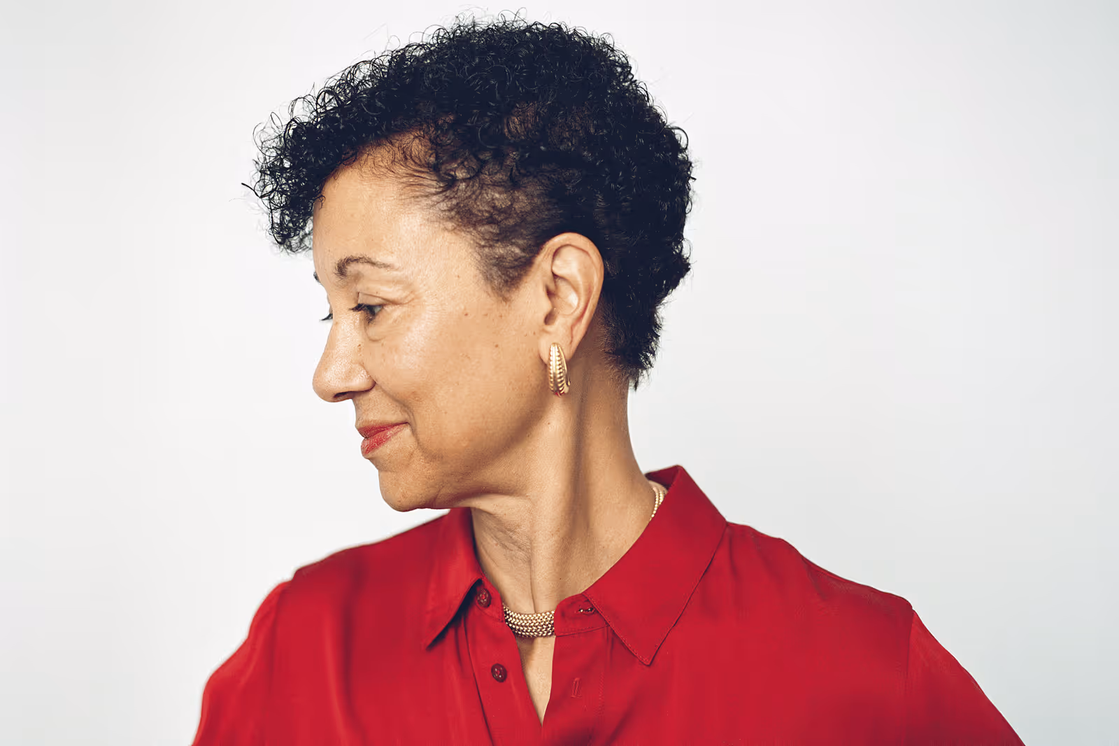 Side profile of Shellye Archambeau looking to the left, wearing a red blouse and gold earrings against a plain white background.