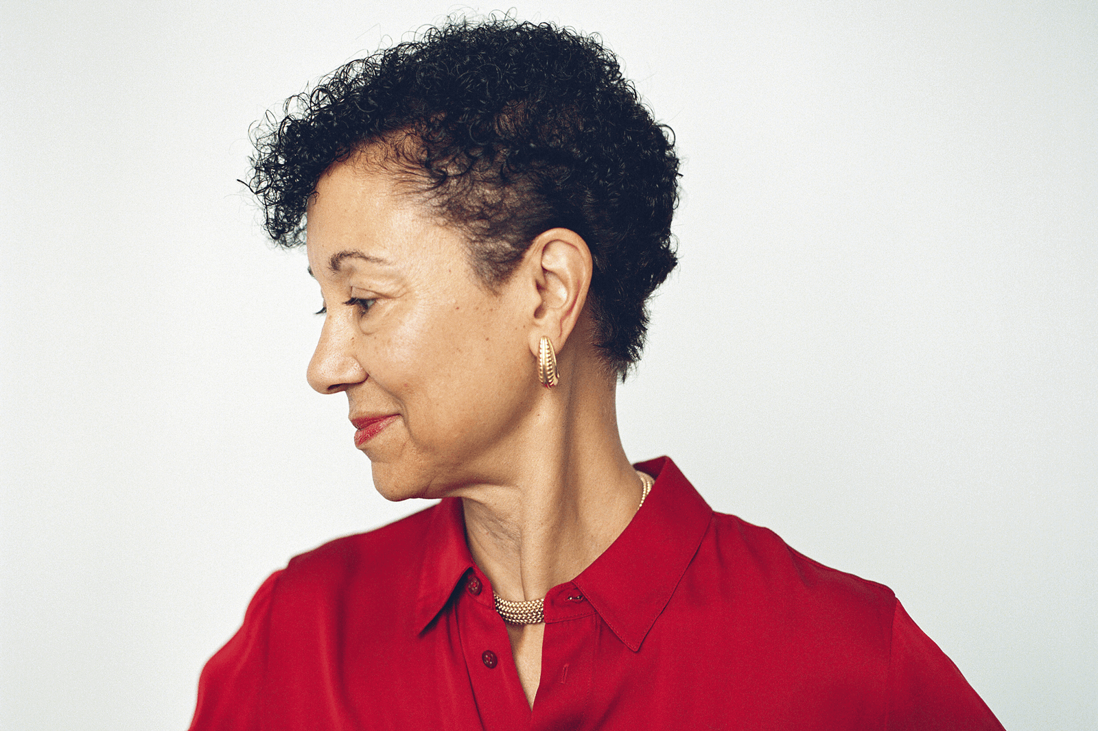 Side profile of Shellye Archambeau looking to the left, wearing a red blouse and gold earrings against a plain white background.