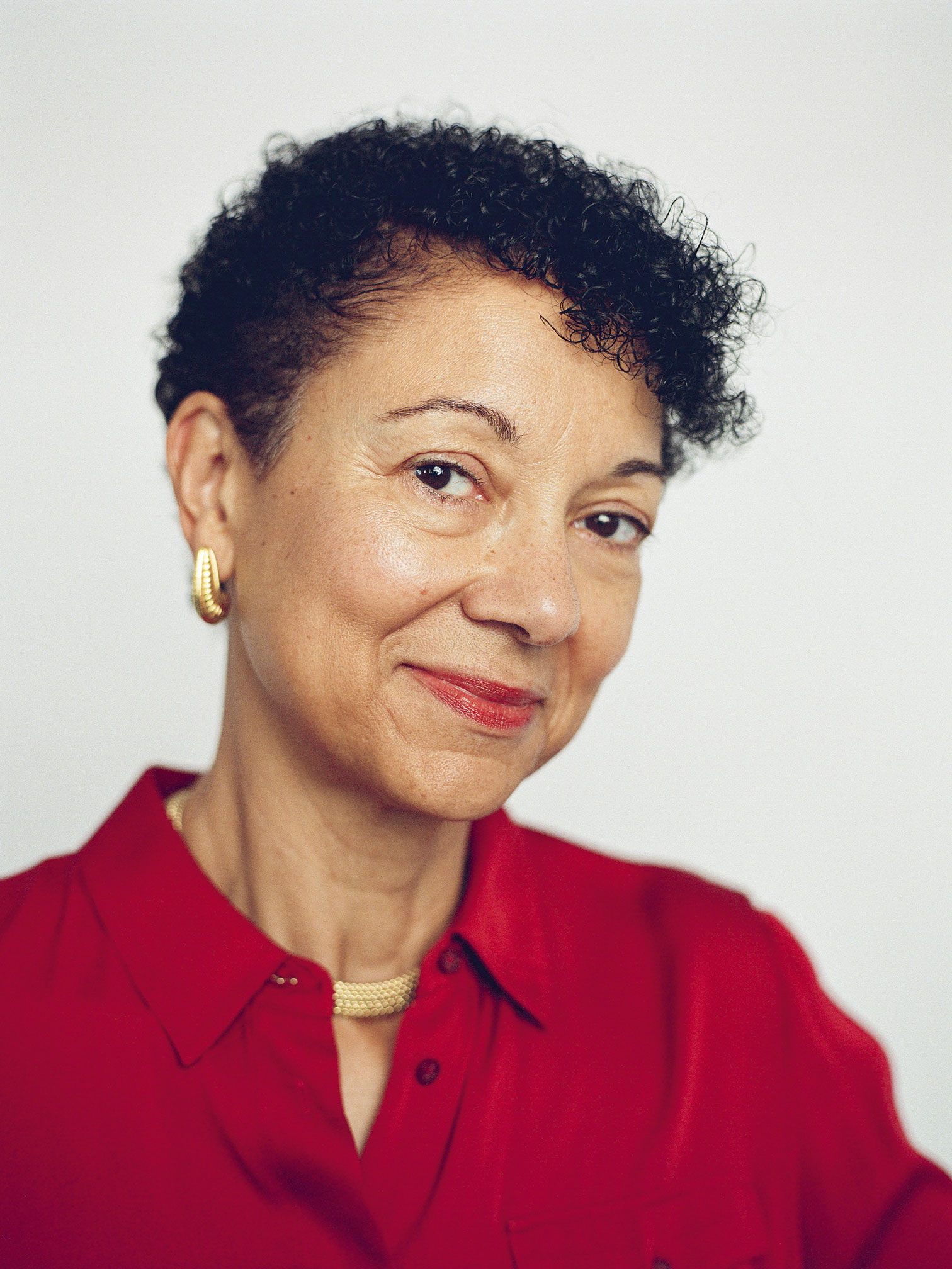 Shellye Archambeau smiles in a red blouse and gold jewelry, shown in a softly lit portrait shot by Ysa Pérez.