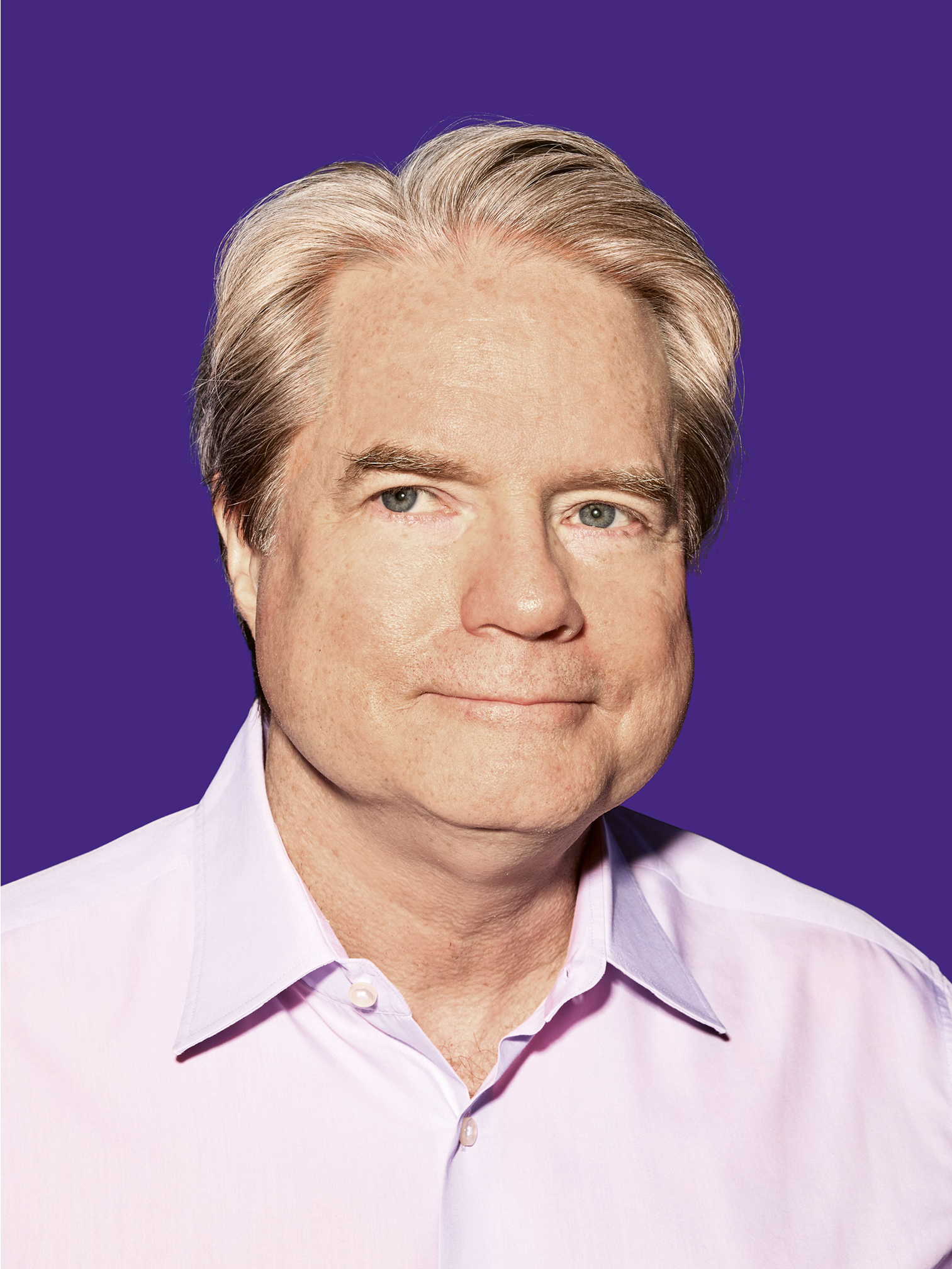 A close up portrait of Michael Morris wearing a purple shirt in front of a purple background, looking slightly sideways to the right.