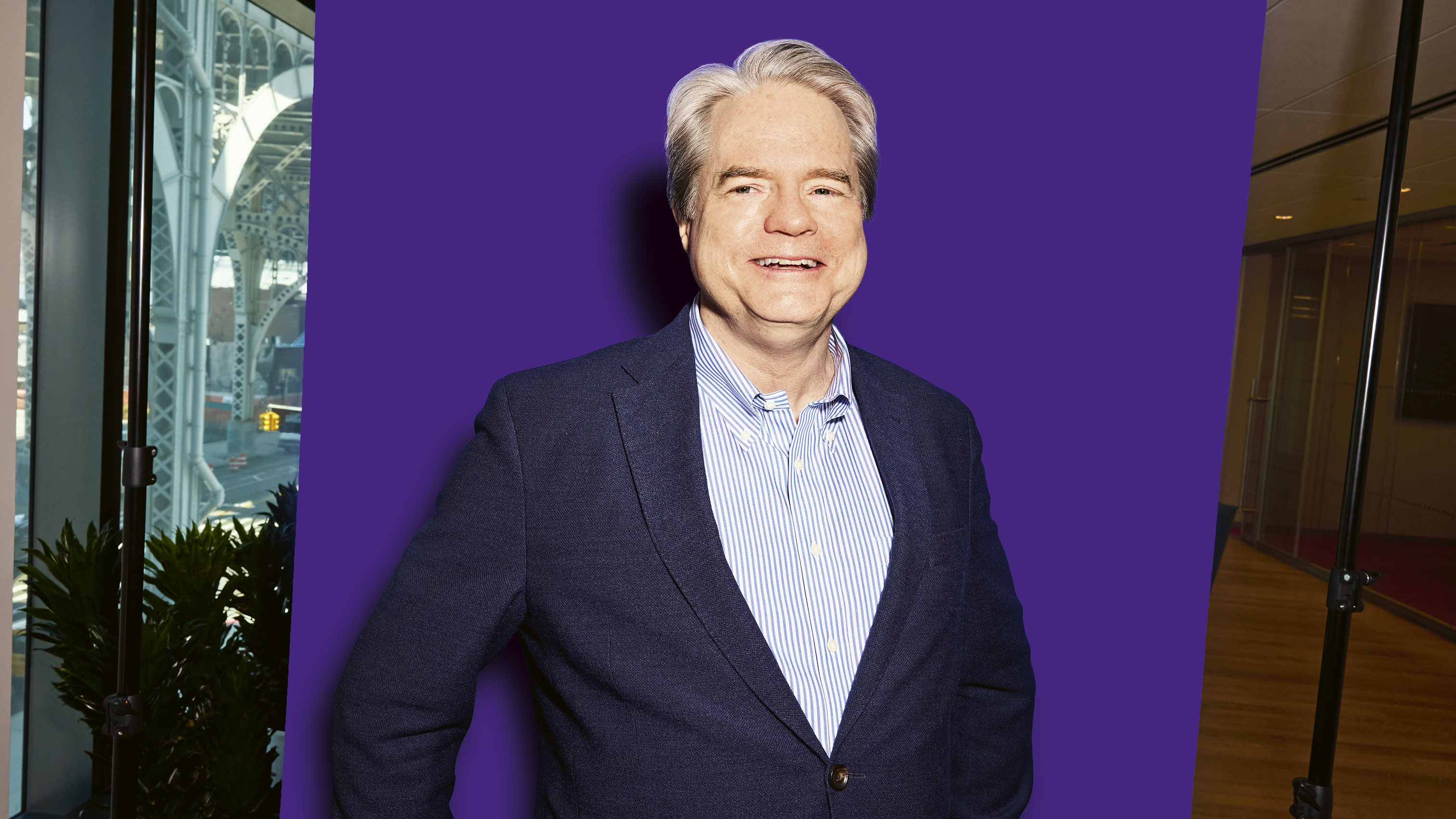 Michael Morris smiling in a blue blazer and striped shirt, standing against a solid purple background with a city scene visible behind glass.