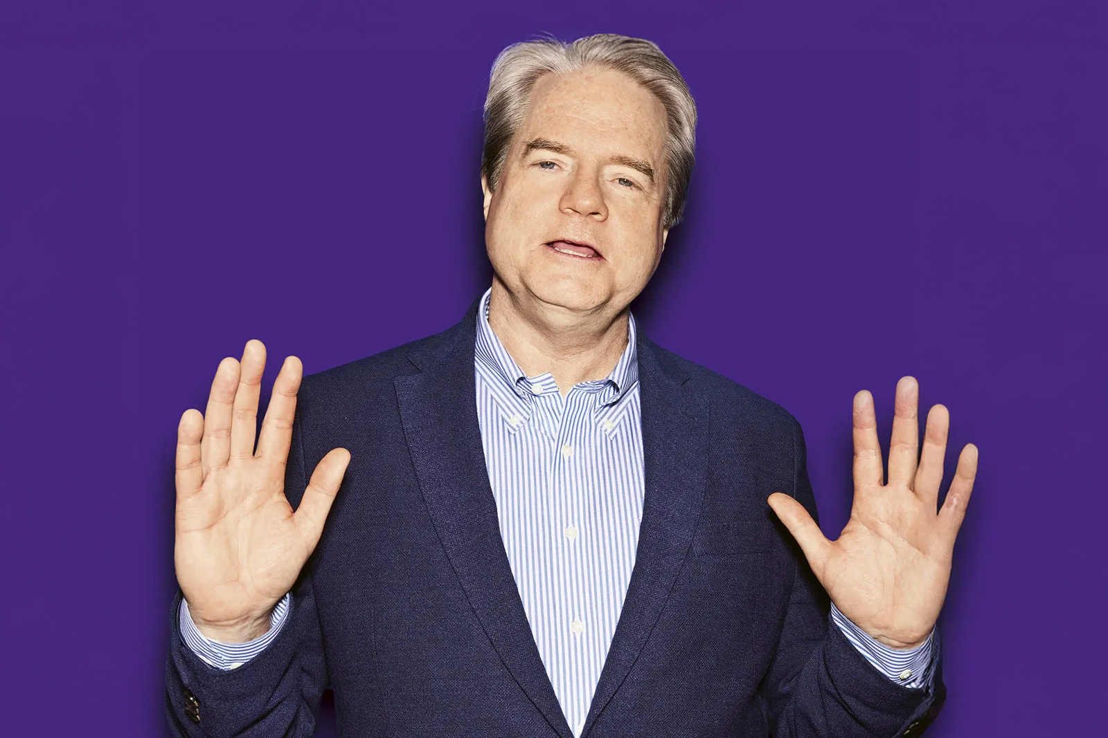 Michael Morris speaking with raised hands, mid-gesture, in a blue blazer and striped shirt against a vibrant purple backdrop.