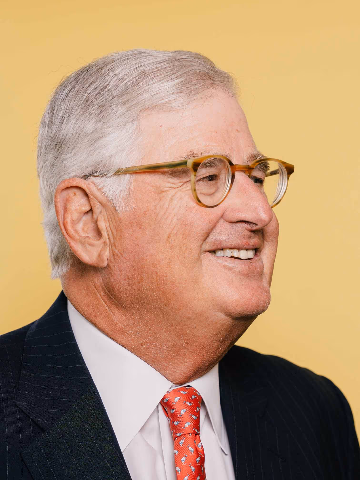 Portrait of Sam Palmisano in a suit and tie, shown in profile against a yellow background. He has short gray hair, wears round glasses and is smiling.