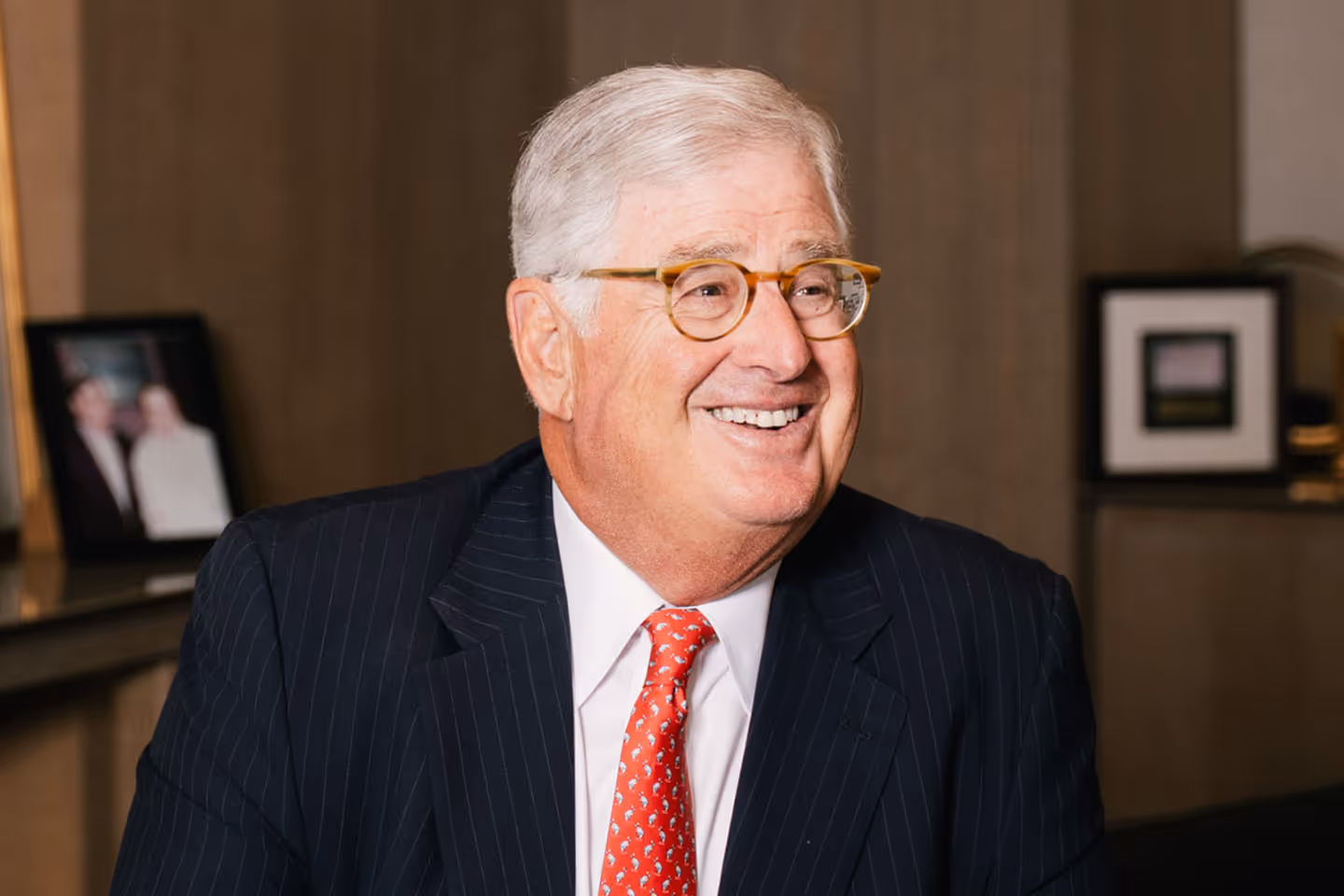 Portrait of Sam Palmisano in a dark pinstripe suit and red patterned tie, smiling while seated in an office setting. He has short gray hair and wears round glasses. Framed photos and office furnishings are visible in the background.