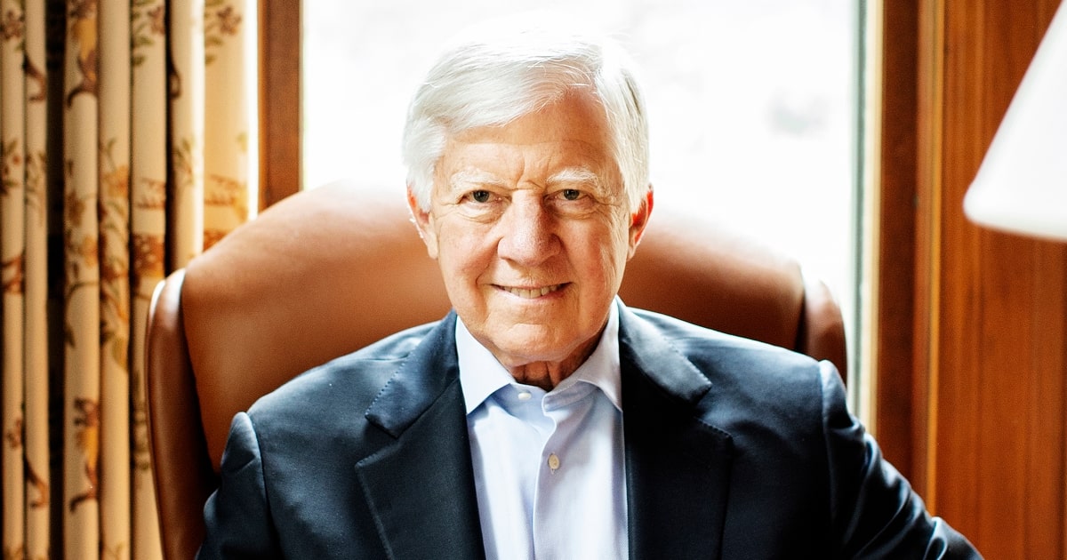 Bill George sits in a leather armchair at a desk, wearing a dark suit jacket and light blue shirt. He faces the camera with a slight smile. Floral curtains and a large window frame the background.
