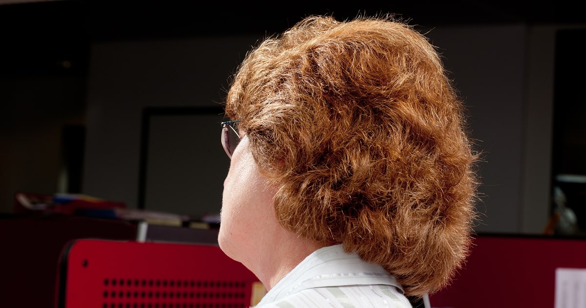 Photograph from Florian van Roekel’s series How Terry Likes His Coffee, showing the back of a person with curly, reddish-brown hair seated at an office workstation. The dimly lit office environment highlights the routines and atmosphere of daily corporate life.