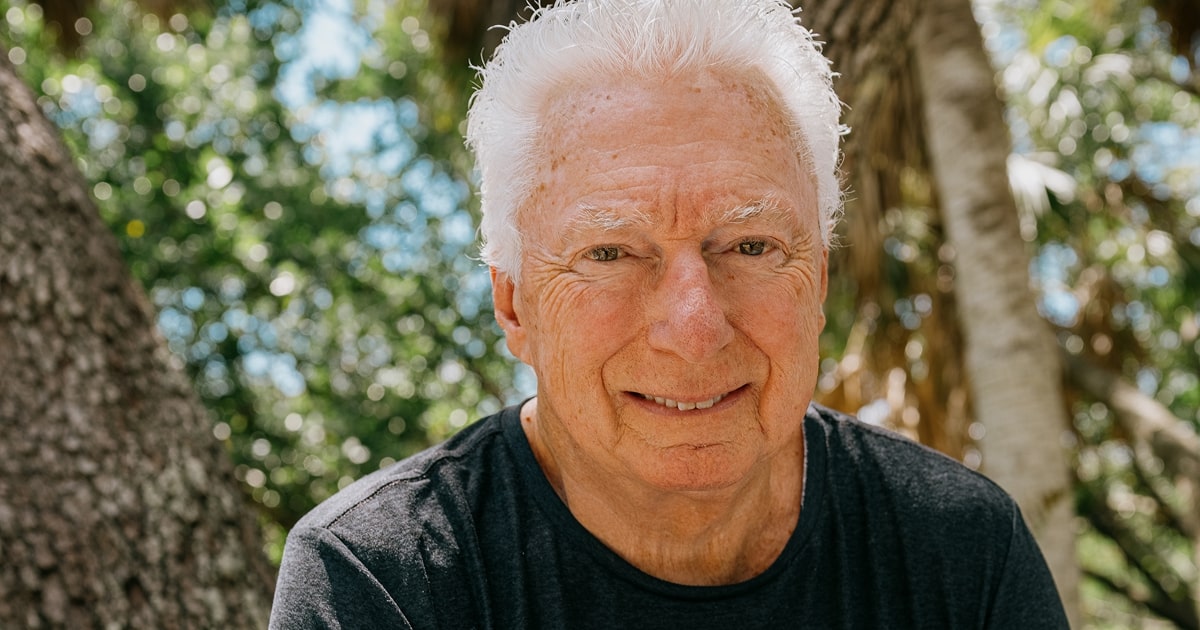 AG Lafley is portrayed with short white hair stands indoors in front of large windows that look out onto a lush, green tropical landscape. He is wearing a black T-shirt and light-colored pants, with one hand resting on a ledge and the other by his side.