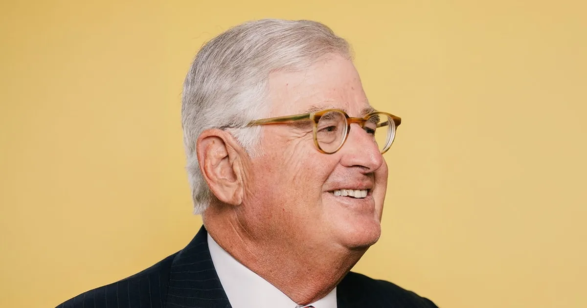 Portrait of Sam Palmisano in a suit and tie, shown in profile against a yellow background. He has short gray hair, wears round glasses and is smiling.