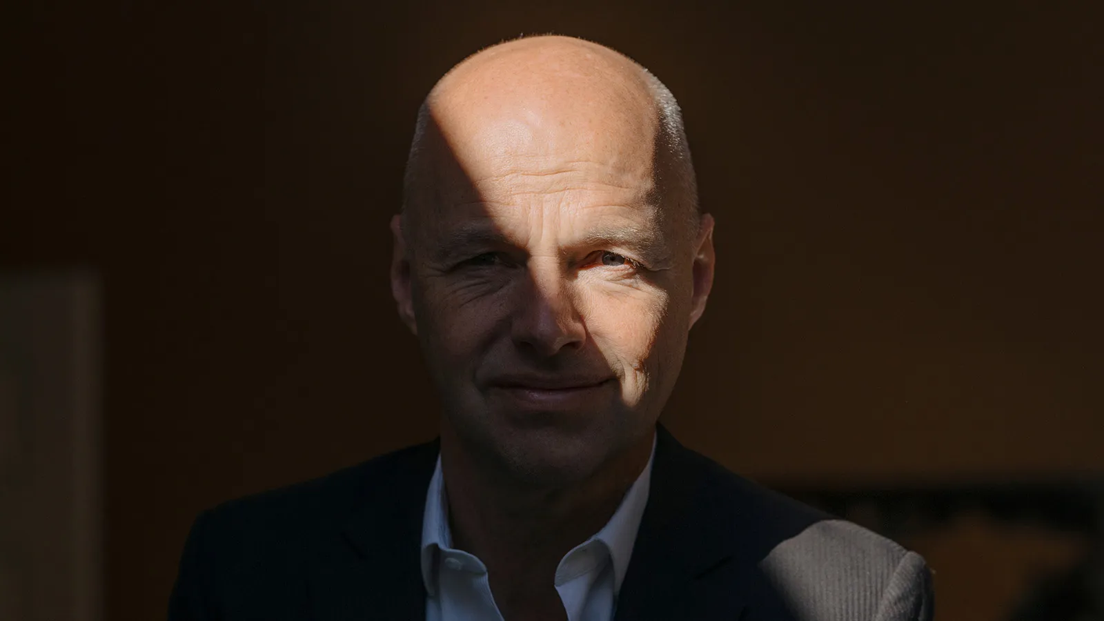 Portrait of Sebastian Thrun in a dark suit and white shirt, seated in dramatic lighting. A diagonal beam of light falls across his face against an otherwise dark background, highlighting his features while the rest of the scene remains in shadow.