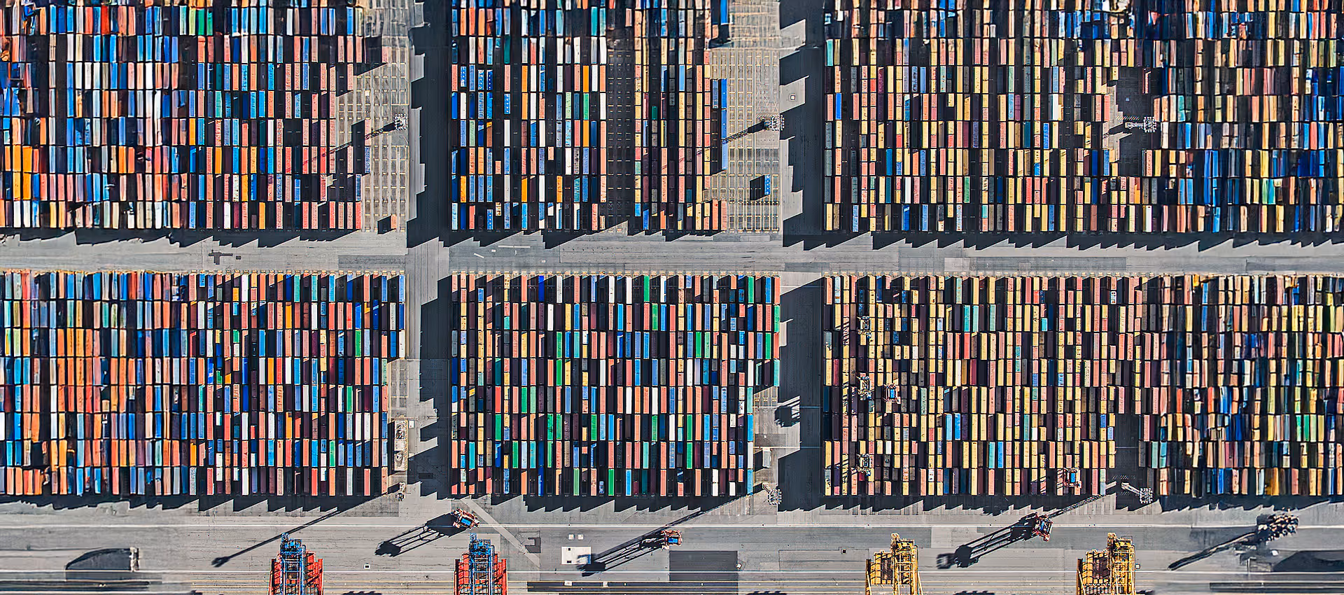 The first image in a series of aerial photograph by Bernhard Lang showing rows of tightly packed, multicolored shipping containers arranged in large rectangular blocks at a port terminal. Trucks and cranes appear at the bottom of the frame, emphasizing the scale of global logistics and containerized trade.