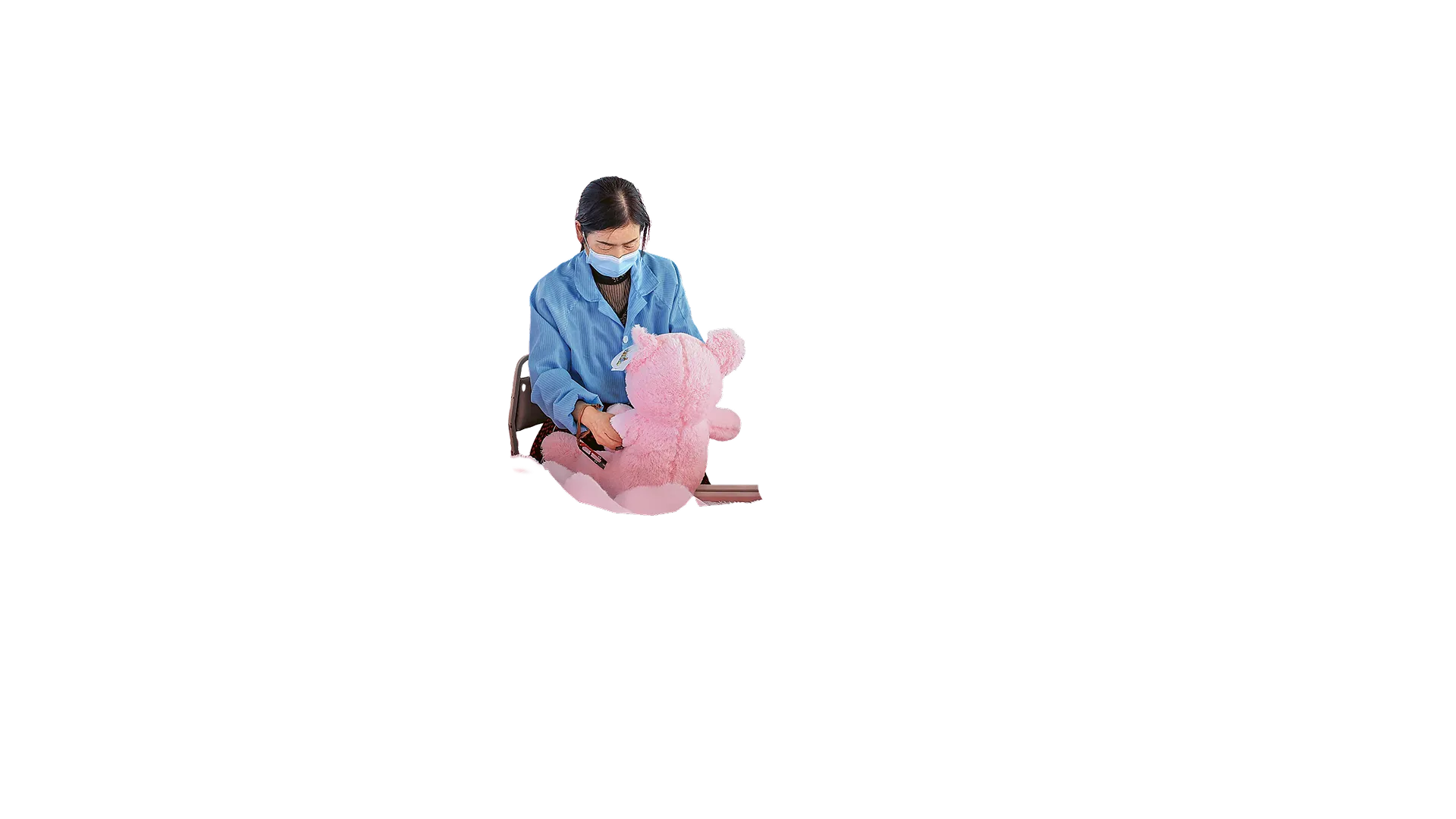 A woman dressed in a blue workers coat is assembling a pink toy. She has black hair and is wearing a face mask. 