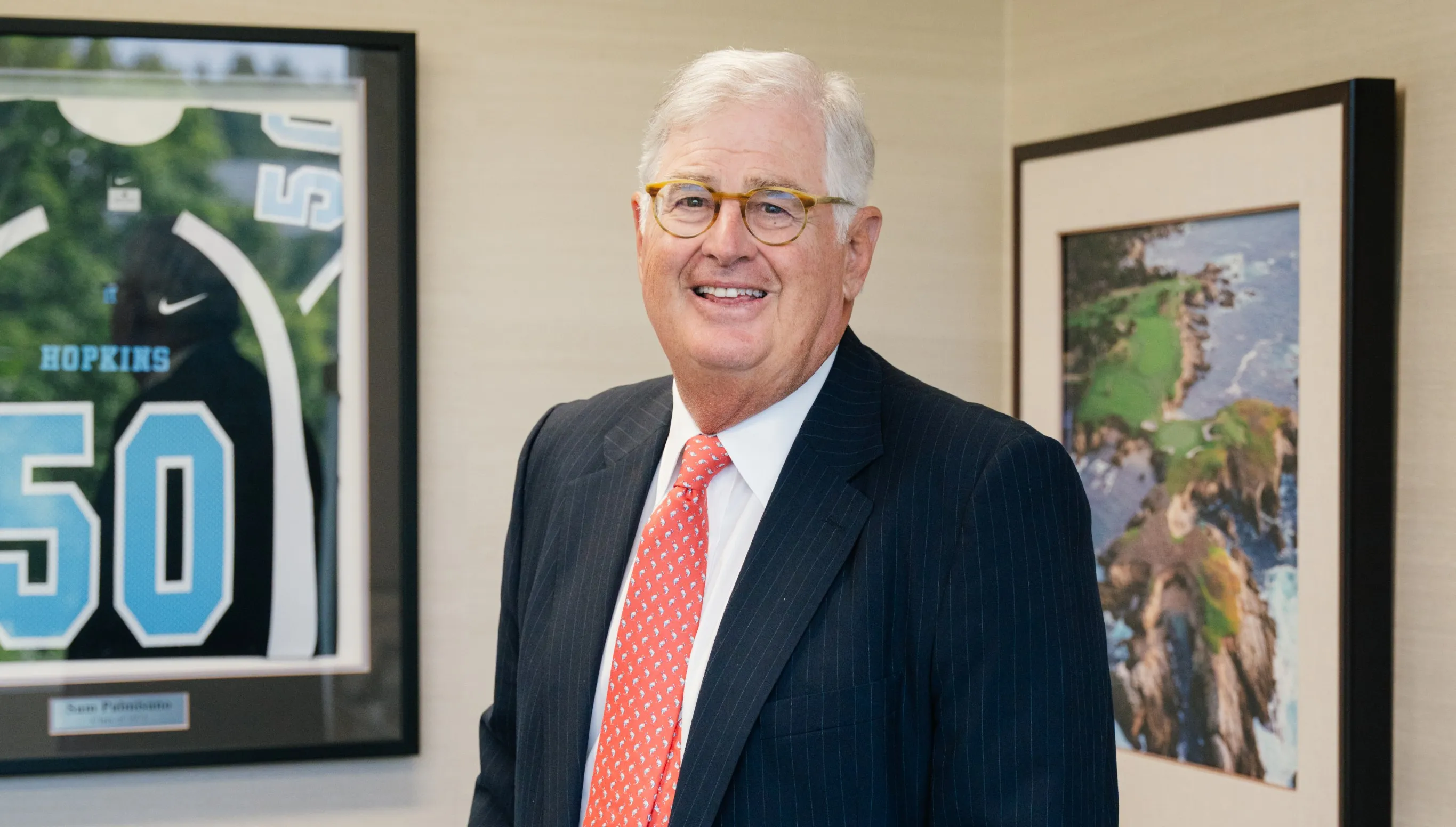 Portrait of Sam Palmisano in a suit and tie, shown his office. He has gray hair, wears round glasses and is smiling. He is standing in front of a framed football jersey with his name and the number “50”, representing his time playing for for the Johns Hopkins football team from 1970-72.