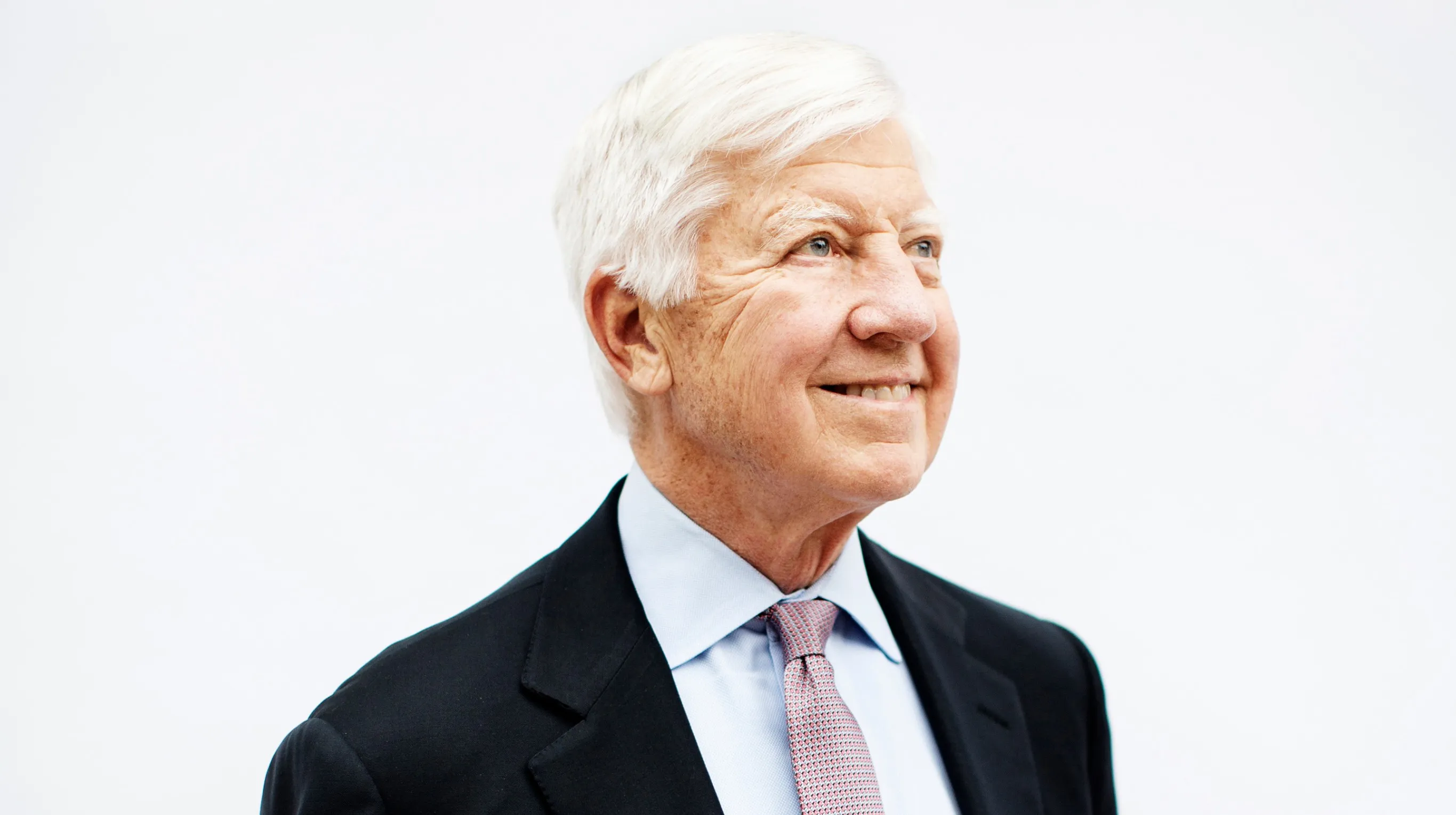 Bill George smiles while looking off to the side. He is wearing a dark suit jacket, a light blue shirt and a textured pink tie, set against a plain white background.