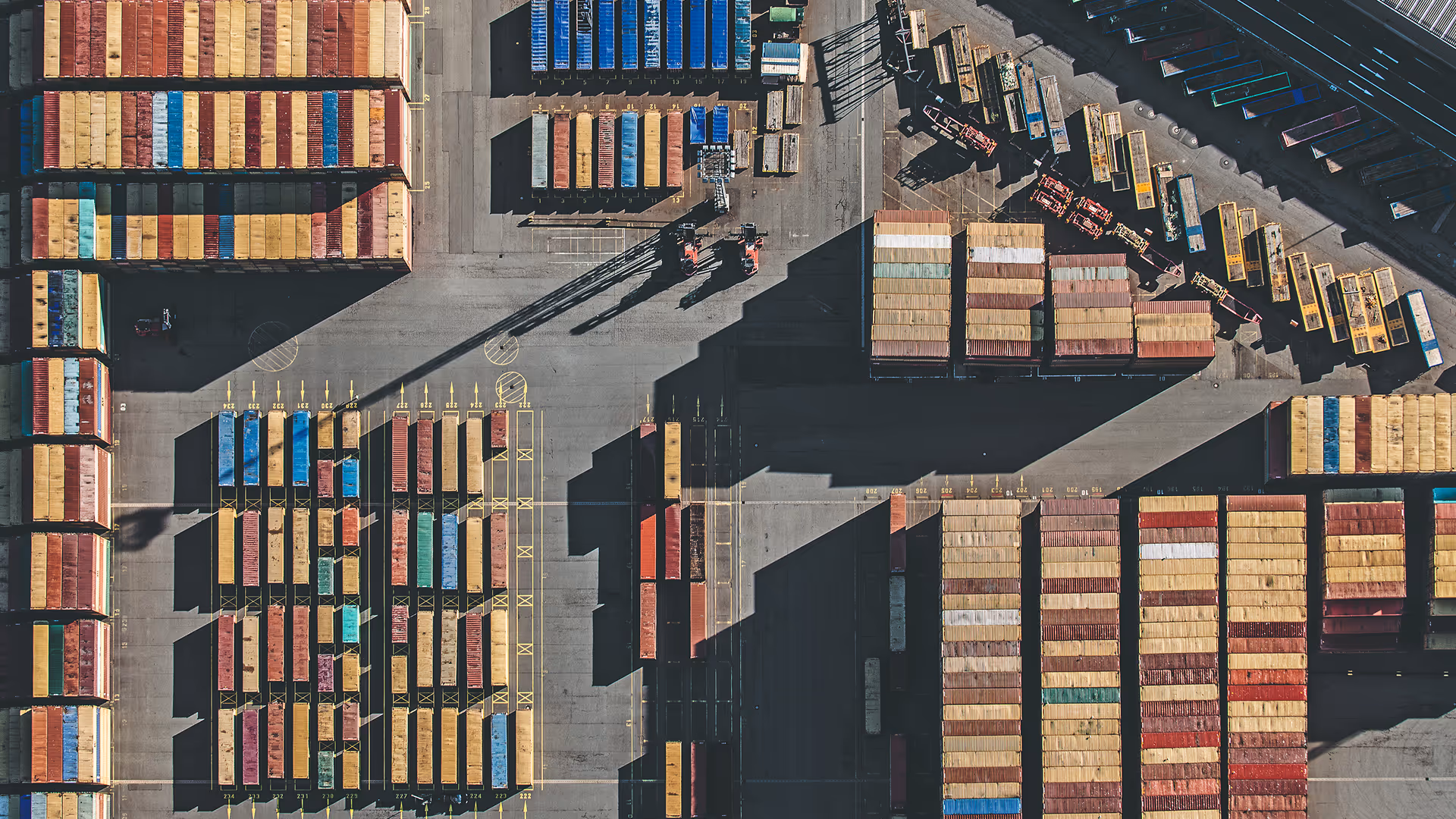 The second aerial photograph by Bernhard Lang showing rows of shipping containers arranged in dense grids across a large port terminal. The image reveals trucks, rail tracks and loading areas, highlighting the scale and infrastructure of global freight and logistics operations.
