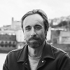 Geoff Poulton looks directly at the camera in a black-and-white portrait. He has a short beard and wears a dark collared jacket over a light t-shirt, standing against a softly blurred background of city buildings.