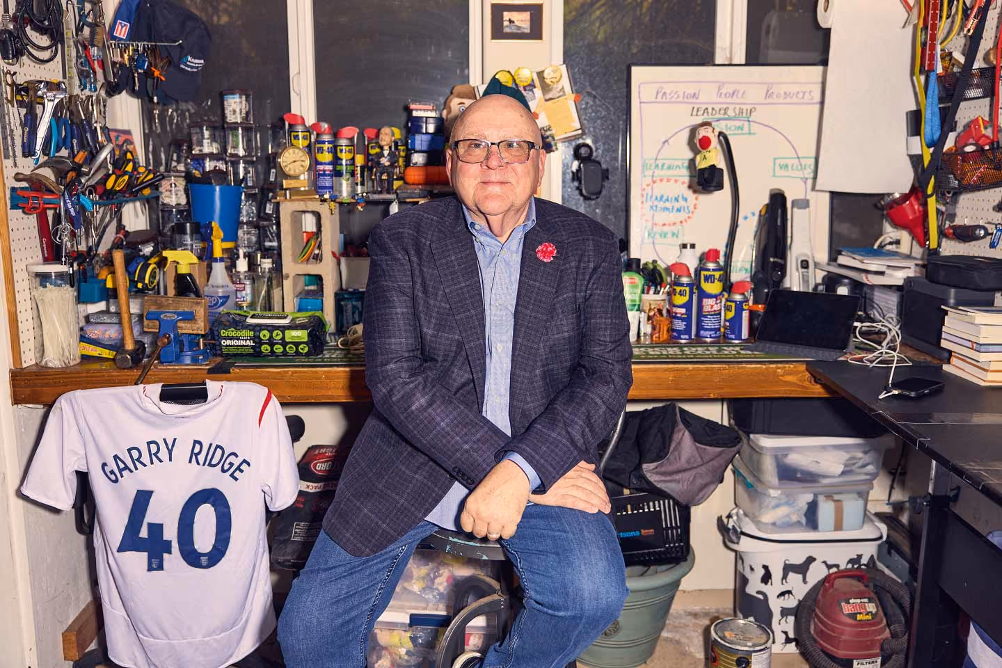 Garry Ridge wears a blazer, and jeans. He sits casually in a highly cluttered workshop. Behind him, a workbench is packed with tools, numerous cans of WD-40, and a whiteboard diagramming leadership concepts. A baseball jersey reading "GARRY RIDGE 40" hangs prominently in the foreground.