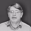 Paul Kielstra looks directly at the camera in a black-and-white portrait, wearing wire-rimmed glasses and a collared shirt against a solid dark background.