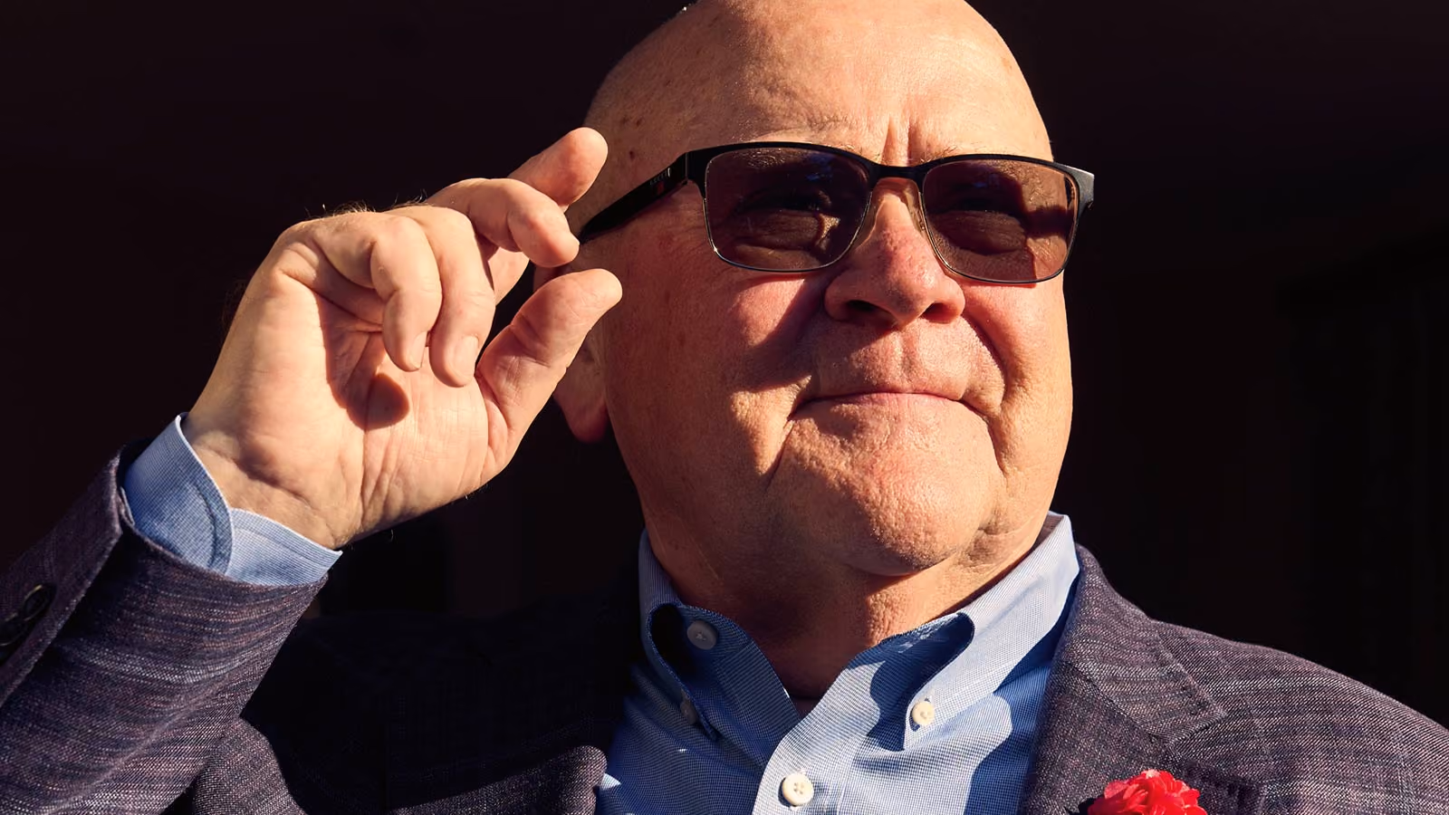 Garry Ridge trägt eine Sonnenbrille, ein hellblaues Hemd und einen strukturierten Sakko mit roter Blumen-Anstecknadel. Er blickt an der Kamera vorbei. Helles, gerichtetes Sonnenlicht beleuchtet seine linke Gesichtshälfte, während er seine rechte Hand vor einem dunklen Hintergrund zu einer dezenten Geste anhebt.