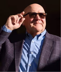 Garry Ridge wears dark sunglasses, a light blue shirt, and a textured blazer with a red floral lapel pin looks off-camera. Bright, directional sunlight illuminates the left side of his face as he raises his right hand in a subtle gesture against a dark background.