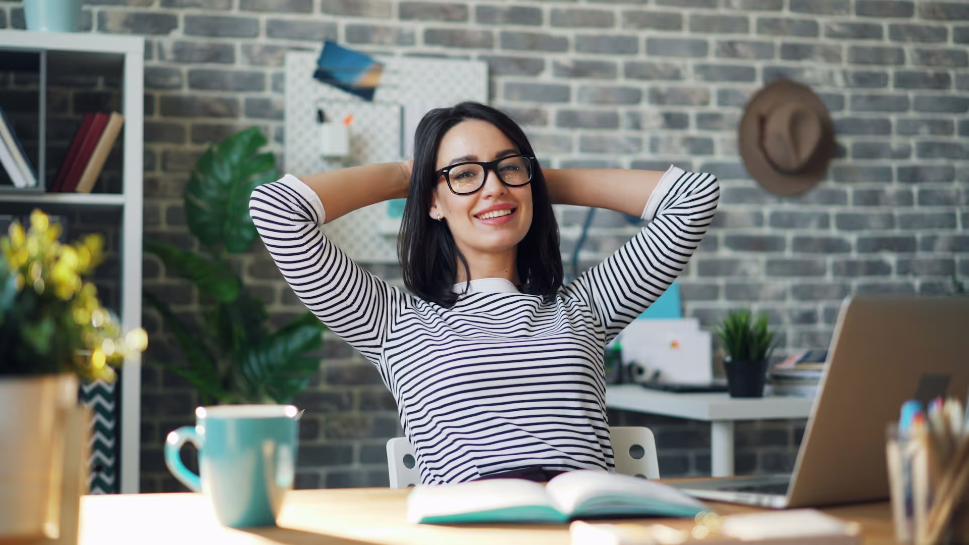 A young woman in her home office, leaning back in her chair in front of her laptop while working.