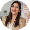 Smiling woman with long dark hair wearing a beige blazer, standing in a modern office setting.