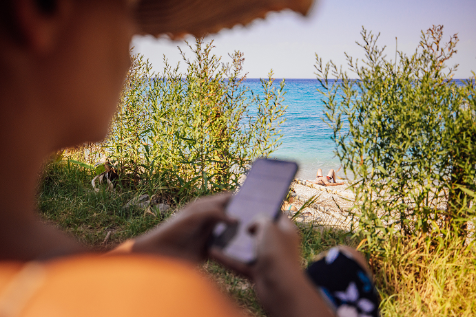 En närbild bakifrån på en person med halmhatt som sitter utomhus i skuggan och tittar på sin smartphone. I bakgrunden, skymd av högt gräs och buskar, syns en vit stenig strand, det turkosa havet och en person som solar.