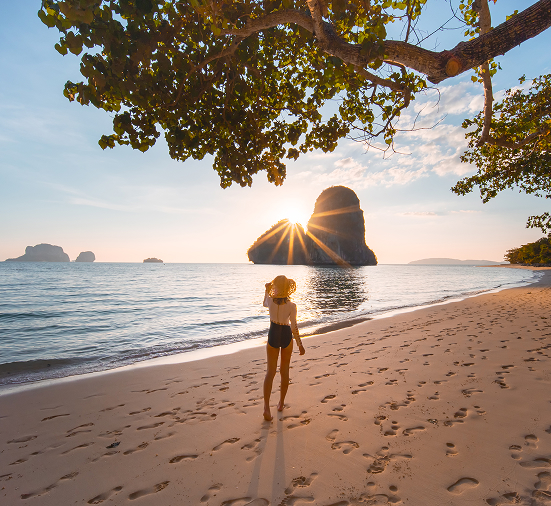 En kvinna i hatt går barfota på en bred, solig sandstrand. Solen står lågt över en stor, karaktäristisk kalkstensklippa som reser sig ur havet i bakgrunden. En stor trädgren hänger ner och ramar in bilden upptill.