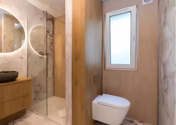 Provence Gaja – Moderne badkamer met inloopdouche, houten kast, ronde spiegel en hangend toilet bij raam.